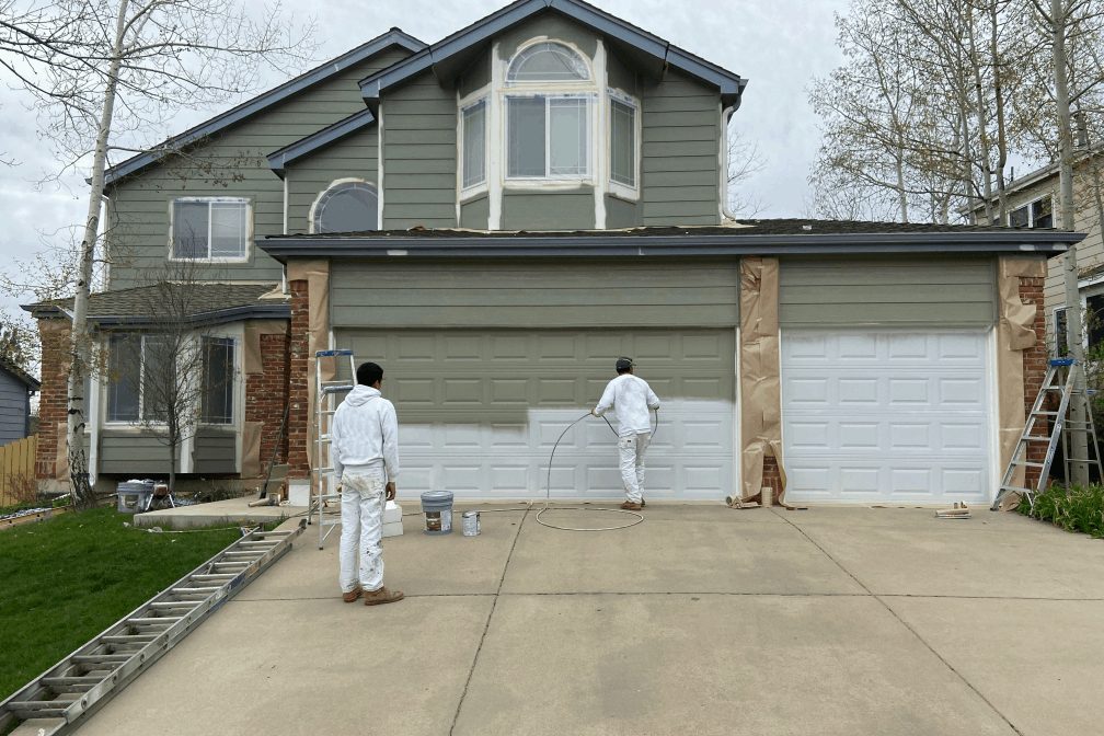 Two painters in white overalls spraying the garage doors of a sage green two-storey house