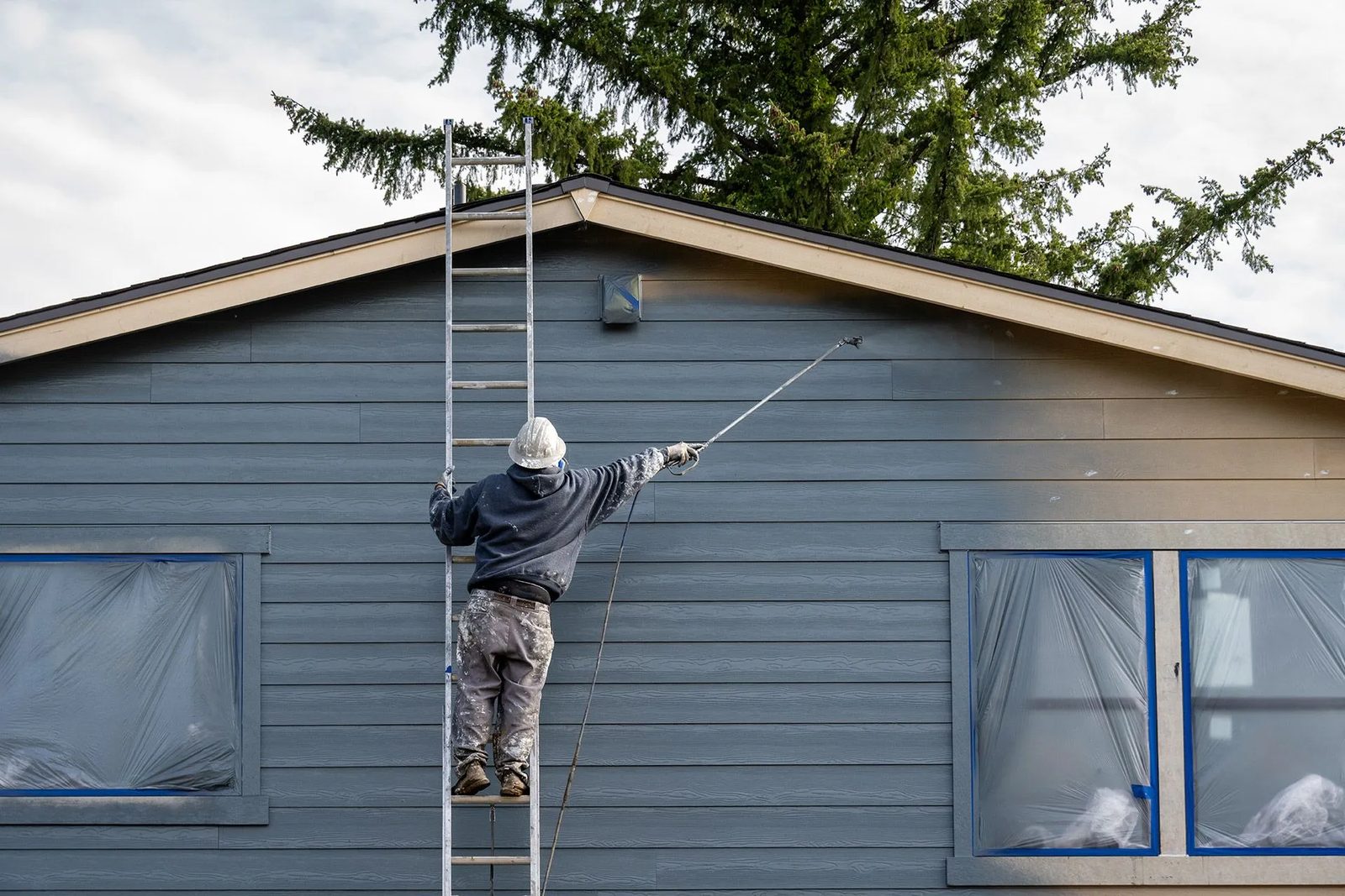 Painter on a ladder spray-painting grey siding with the windows masked off